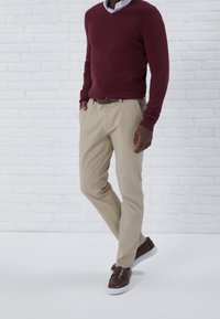 Maglione borgogna con scollo a V, abbinato a pantaloni color khaki. Scarpe brown senza lacci, in piedi su un pavimento bianco contro uno sfondo di muro di mattoni.