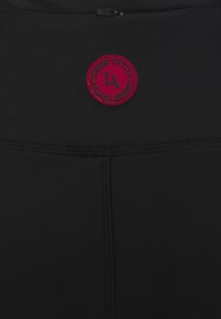 Melnas sporta apģērbs ar strukturētu audumu, kas piedāvā apaļu sarkanu logo uzlīmi. Iekļauta rāvējslēdzēja kabata. Gludās šuves uzlabo dizainu.