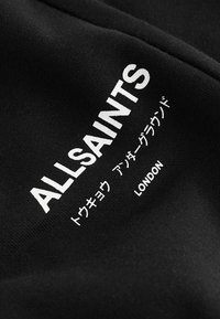 Melnas audums ar baltiem burtiem. "ALLSAINTS" treknrakstā, kopā ar japāņu rakstzīmēm un "LONDON" mazākā fontā apakšā. Teksts ir centrāli izvietots.