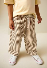 Kleinkind trägt beige-weiße, vertikal gestreifte Hosen mit Kordelzug an der Taille und weiße Sneakers mit roten und blauen Streifen.