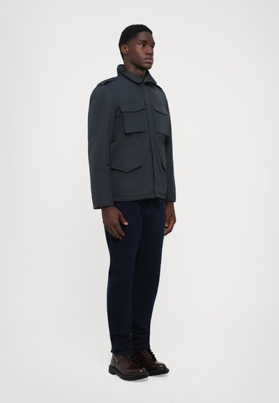 Chaqueta azul marino con cremallera, cuatro grandes bolsillos frontales y cuello. Lleva pantalones oscuros y botas marrones; con un fondo sencillo.