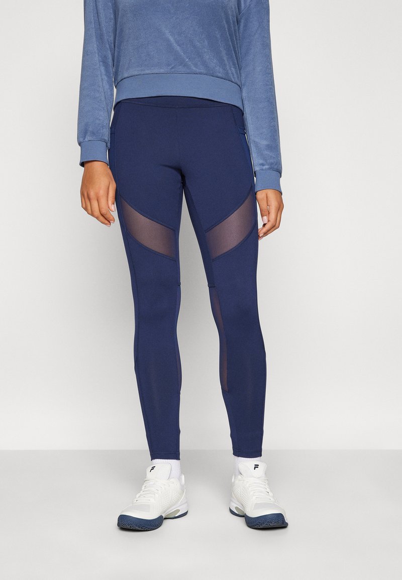 Leggings blu navy con pannelli in mesh trasparente sui lati, abbinati a un maglione in pile blu e scarpe sportive bianche. Tessuto morbido e design aderente.