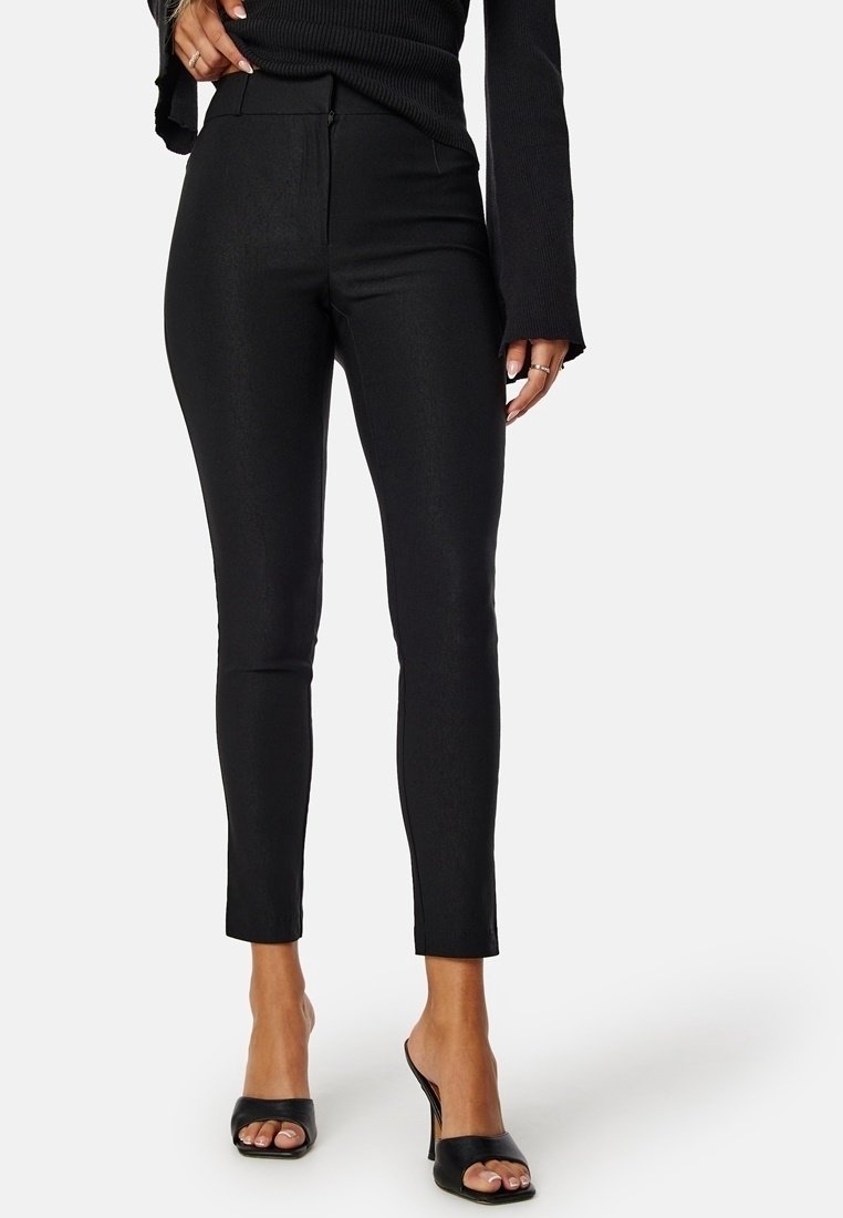 Pantalon skinny noir à texture lisse, taille mi-haute et longueur cheville. Associé à des mules noires à bouts ouverts. Aucun motif ou accent visible.