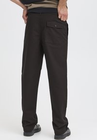 Musta, kevyet housut rentolla istuvuudella, joissa on takatasku napillinen suljin ja teksturoitu kangas, nähtynä takaa.