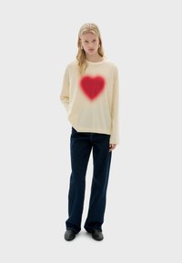 Beige trui met een rood hartontwerp in het midden, lange mouwen, gecombineerd met donkerblauwe wijde jeans en zwarte schoenen.
