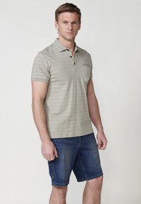 Khaki pruhované polo triko s přední kapsou, límcem na tři knoflíky a krátkými rukávy, v kombinaci s džínovými šortkami, stojící na jednolitém pozadí.