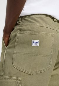 Pantalons de travail Lee en vert olive avec une grande poche arrière et une étiquette blanche. Le tissu présente une texture lisse avec des détails de couture visibles.