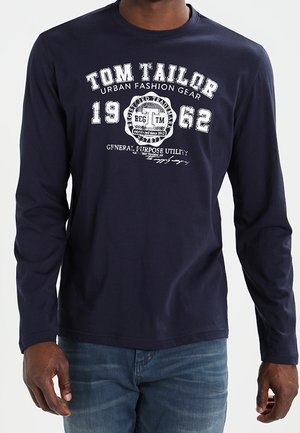 Mężczyzna w granatowej koszuli z długim rękawem z białym napisem "Tom Tailor Urban Fashion Gear 1962" oraz w niebieskich dżinsach.