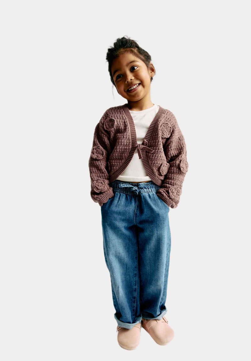 Stickad brun kofta med blommönster, vit skjorta, lösa blå denimbyxor och ljusrosa skor. Modellen står med händerna i fickorna.