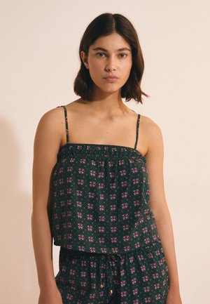 Młoda kobieta z krótkimi ciemnymi włosami, ubrana w zielony bezrękawnik w kwiatowy wzór i dopasowane spodnie na jednolitym beżowym tle.