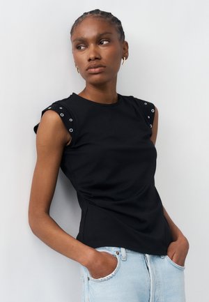 Νεαρή γυναίκα με πλεξούδες, φοράει μαύρο αμάνικο τοπ με διακοσμητικές λεπτομέρειες, ανοιχτόχρωμο μπλε τζιν, στέκεται με τα χέρια στις τσέπες.