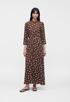 Femme portant une robe longue marron à manches longues boutonnée avec des pois blancs et des tongs noires, debout devant un fond blanc uni.