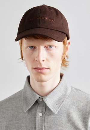Un bonnet marron avec le logo "HOLZWEILER" brodé en couleur rouille. La personne porte une chemise grise à col. Gros plan, fond neutre, expression concentrée.