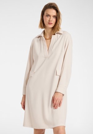 Robe beige à manches longues avec un col, décolleté en V et deux poches frontales. Fabriquée dans un tissu lisse avec une coupe décontractée.
