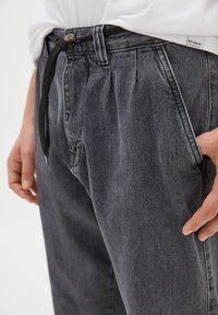 Šedé denimové kalhoty mají volný střih s šňůrkovým pasem a záhyby vpředu. Textura látky vypadá měkce a mírně opotřebovaně.