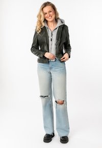 Zwarte leren jas over een grijze hoodie, gecombineerd met lichtblauwe gescheurde jeans en zwarte stevige laarzen. Model staat met een glimlach.