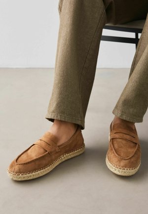 Braune Wildleder-Slipper mit geflochtenen Jutesohlen, getragen zu olivgrünen Hosen, an einer Person, die vor einem neutralen Hintergrund sitzt.