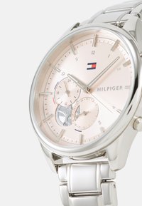 Sølv rustfritt stålklokke med en lys rosa skive, store timemarkører og sub-dials. Har en logo og teksturerte aksenter på klokkens ansikt.