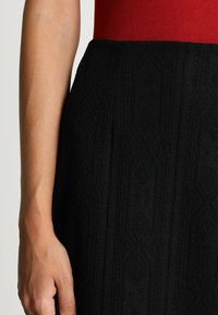 Jupe noire texturée avec un design à motifs, associée à un haut rouge ajusté. Gros plan sur une main reposant sur la taille.