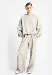 Kobieta w beżowym, oversize'owym hoodies i szerokich spodniach, stojąca z rękami w kieszeniach, na tle białym.