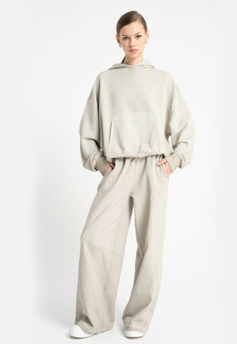 Kobieta w beżowym, oversize'owym hoodies i szerokich spodniach, stojąca z rękami w kieszeniach, na tle białym.