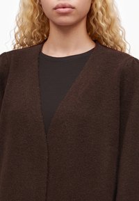 Femme aux cheveux blonds bouclés portant un cardigan marron texturé sur un t-shirt noir à col rond, montrée des épaules aux lèvres.