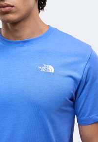 Άνδρας που φοράει μπλε T-shirt της The North Face με το λογότυπο στο αριστερό πάνω μέρος του στήθους, με απλό φόντο.