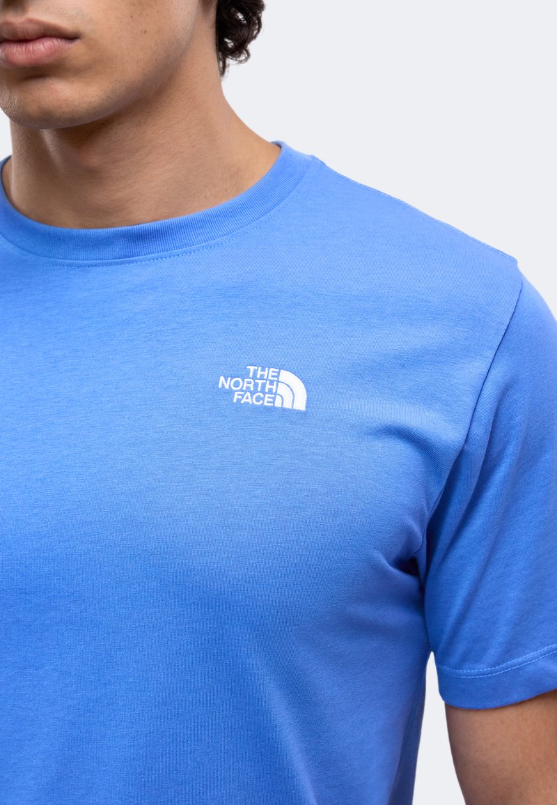 Hombre con camiseta azul de The North Face con logo en la parte superior izquierda del pecho sobre un fondo liso.