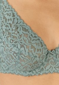 Sostenes de encaje en un tono verde azulado suave con un patrón floral, bordes en forma de onda y un suave forro. Presenta un escote pronunciado y un delicado lazo como acento.