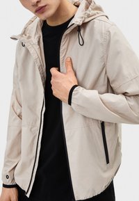 Ljusbeige jacka i lätt tyg med huva, dragkedja framtill, elastiska muddar och svarta detaljer på dragkedjan och fållen.