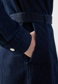 Dark denim jacket with a belted waist, featuring a button cuff and side pockets, textured fabric with subtle stitching details.