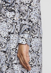 Camisa de manga longa com um fundo preto decorado com intrincados padrões florais paisley em azul e branco, com punhos abotoados e textura de tecido suave.