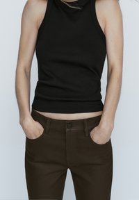 Vrouw in een mouwloos zwart topje, gestoken in een donkerbruine broek met handen in de zakken tegen een simpele achtergrond.