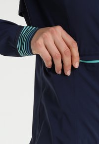 Veste marine avec poignet rayé bleu sarcelle. Présente un design élégant, une texture lisse et une petite poche avec un accent bleu sarcelle le long de la couture.