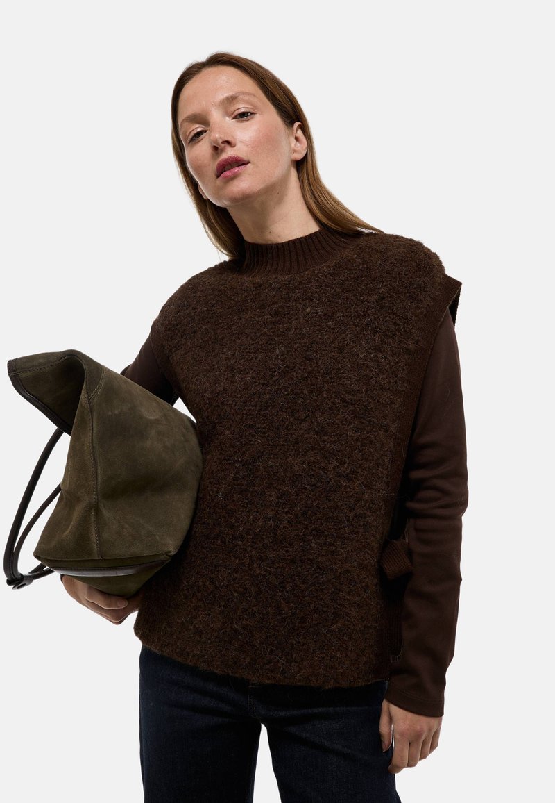 Pull en couches marron avec une surface texturée et duveteuse, col haut et manches courtes, associé à un sac en cuir suédé vert. Un jean foncé complète le look.