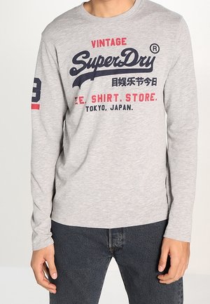 Person iført en lysegrå langærmet skjorte med "Vintage SuperDry" og japanske tegn trykt på fronten, kombineret med mørke jeans.