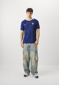 Maglia blu a maniche corte, colletto bianco e emblema francese; indossata con pantaloni cargo in denim chiaro e larghi e sneakers bianche.