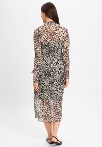 Durchsichtiges, langärmliges Midikleid mit einem schwarz-beigen gepunkteten Muster. Geraffte Taille und fließende Textur, kombiniert mit schwarzen flachen Sandalen.