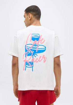 Homme de dos portant un t-shirt blanc avec un motif de moto bleu et un texte rose « Cafe Racer » dans le dos, associé à un short rouge.