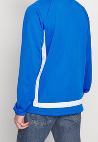 Sininen collegetakki pyöreällä kauluksella, valkoiset sivupaneelikoristeet, pitkät hihat ja pehmeä materiaali. Kaksi sivutaskua alaosassa.