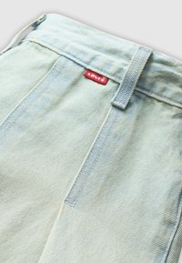 Gros plan sur un tissu en denim bleu clair montrant la ceinture, la boucle de ceinture, les coutures et une petite étiquette rouge de la marque Levi's.
