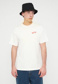 Άσπρο βαμβακερό μπλουζάκι με ένα μικρό κόκκινο γραφικό σκέιτμπορντ στην αριστερή πλευρά του στήθους, συνδυασμένο με μπλε τζιν και μαύρο καπέλο τύπου κάδο.