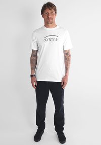 Homme avec des tatouages sur les deux bras portant un t-shirt blanc OXBOW, un pantalon noir, des sneakers noires et une montre noire, se tenant devant un fond uni.