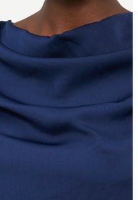 Κοντινό πλάνο σκούρου δέρματος στον λαιμό και το άνω μέρος του στήθους, που φοράει ένα ζεστό μπλε ύφασμα με απαλές πτυχώσεις και λαιμόκοψη λίγο εκτός ώμου.