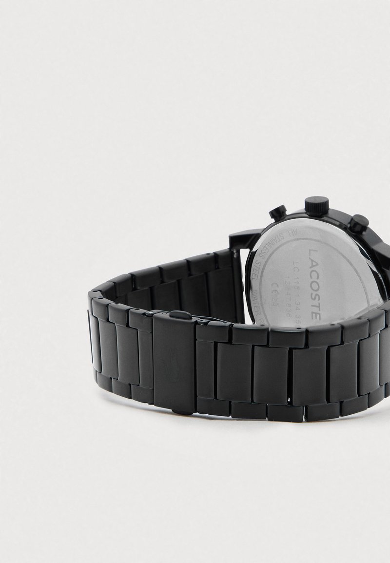 Zwart metalen linkarmbandhorloge dat de achterkant van de kast toont met de merknaam Lacoste en details over waterbestendigheid.