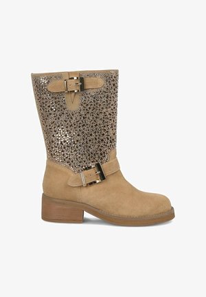 Beżowe zamszowe botki z niskim słupkowym obcasem, błyszcząca zdobiona cholewka oraz dekoracyjna klamra na pasku wokół kostki.