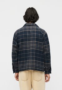 Άτομο που φοράει σκούρο μπλε και μπεζ καρό σακάκι και μπεζ παντελόνι, στέκεται με την πλάτη προς τα εμπρός σε απλό λευκό φόντο.