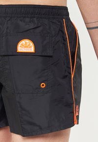 Costumi da bagno neri con fascia elastica in vita, cuciture di accento arancioni, una tasca sul retro con una patch arancione "SUNDEK" e un dettaglio con occhiello.