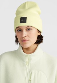 Zelený pruhovaný beanie v limetkové barvě s černou logovou nášivkou, nošený s měkkou světle zelenou fleecovou bundou se zipem a strukturovanou tkaninou.