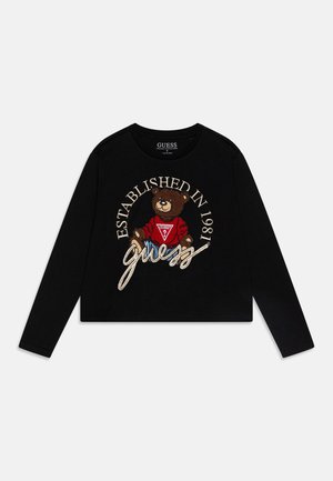 Sort langærmet skjorte med en bamse iført en rød sweater, tekst "Established in 1981" og kursiv "guess" logo foran.